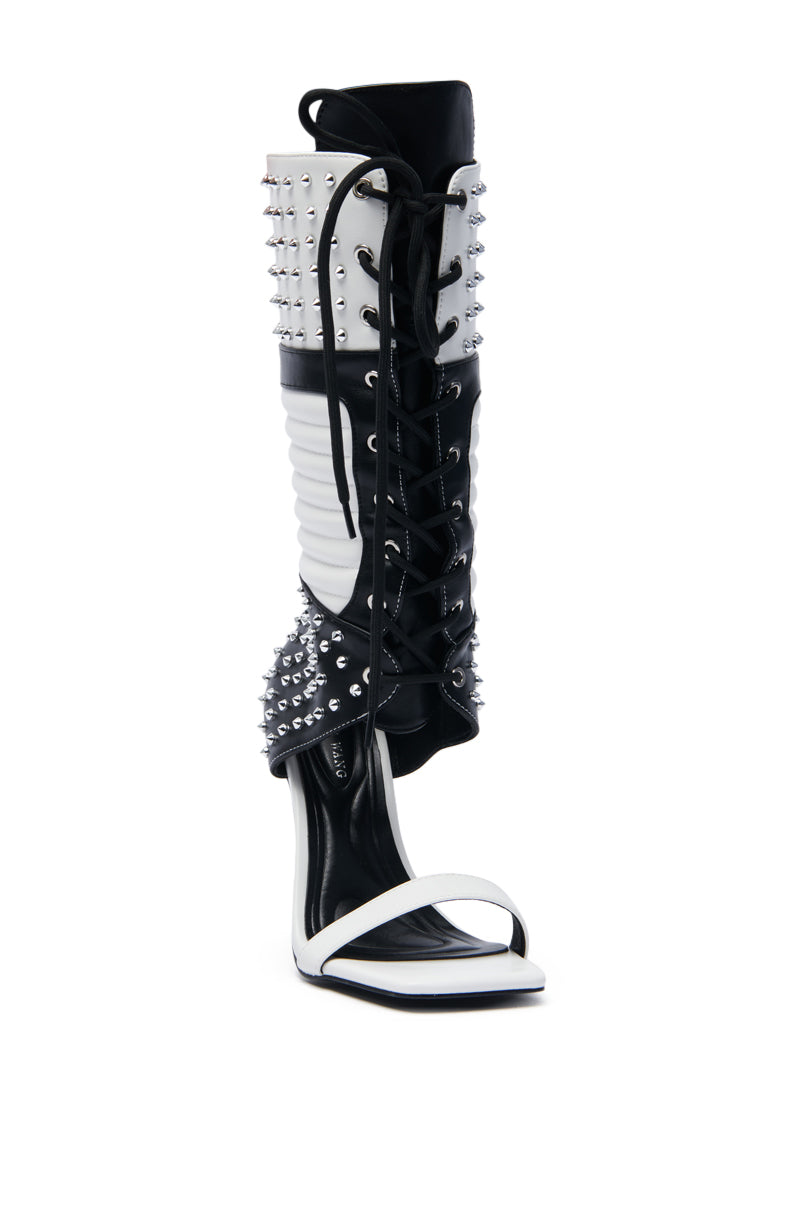 The AZALEA WANG ALBERTO BLACK WHITE STUDDED OPEN TOE BOOT is a high-heeled, open-toe boot with black and white faux leather, silver studs, lace-up front, and intricate cut-out details on the stiletto heel and ankle.