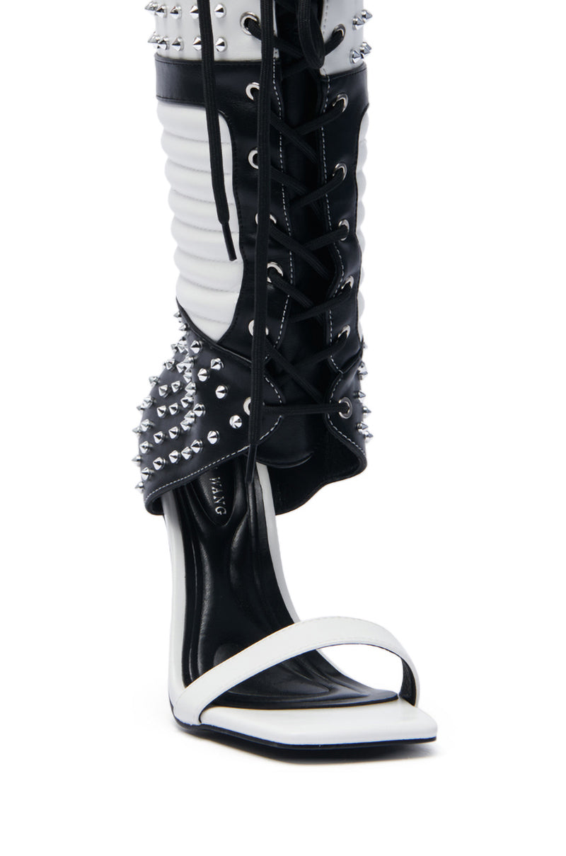 The AZALEA WANG ALBERTO BLACK WHITE STUDDED OPEN TOE BOOT showcases a chic stiletto heel with a black and white design, featuring a white open toe strap, faux leather lace-up front, and an ankle section adorned with silver studs. Its slender black heel completes the edgy look.