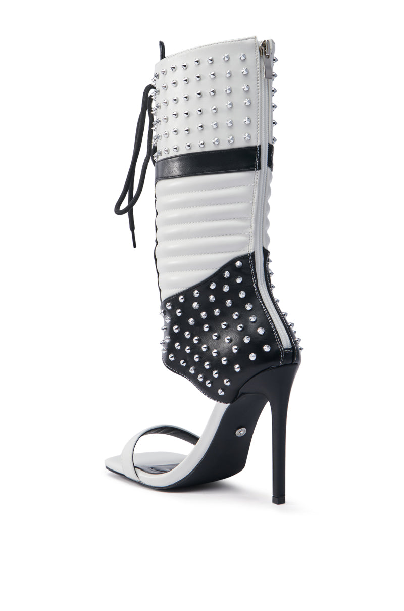 The AZALEA WANG ALBERTO WHITE AND BLACK GLADIATOR SPORTY BOOT is a high-heeled boot with a bold white and black design. It's made of faux leather adorned with silver studs, featuring a stiletto heel, side zipper, and black lace-up detail at the top for an edgy twist.