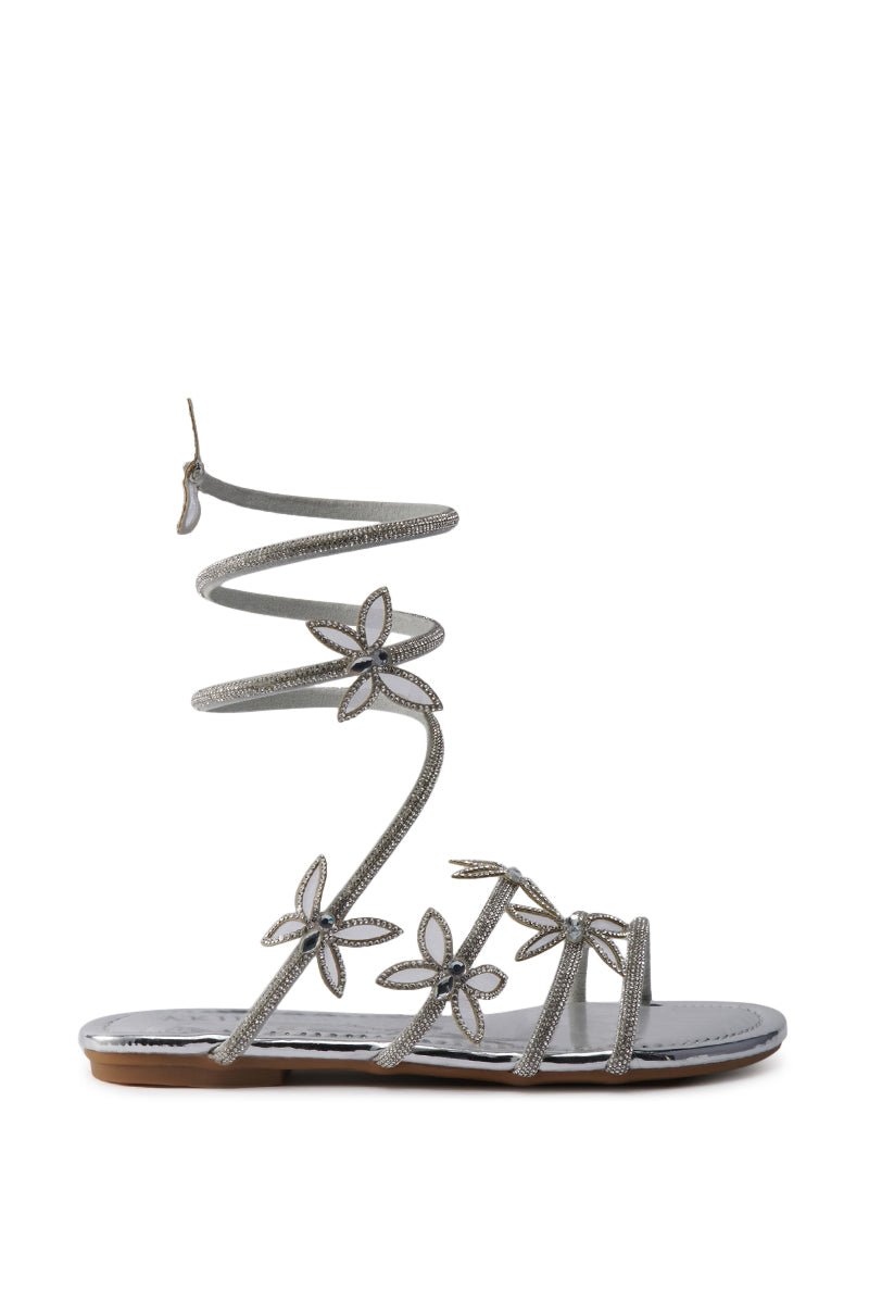 The AKIRA ALBUS SILVER COIL RHINESTONE FLAT SANDAL features metallic faux leather, a wraparound ankle strap, and rhinestone-studded floral accents on slim straps, shown against a white background. 3