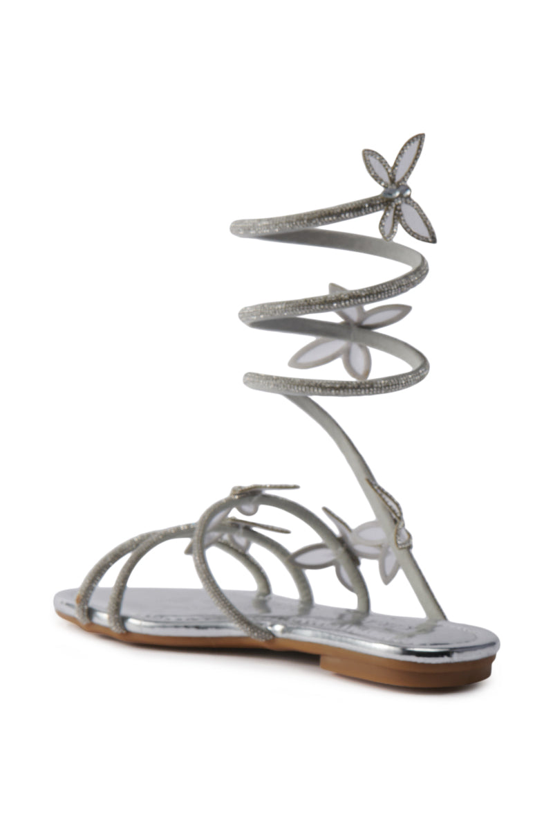 The AKIRA ALBUS SILVER COIL RHINESTONE FLAT SANDAL features metallic faux leather, a spiral ankle-wrap strap embellished with rhinestones, and decorative flower accents on both the straps and spiral. 5