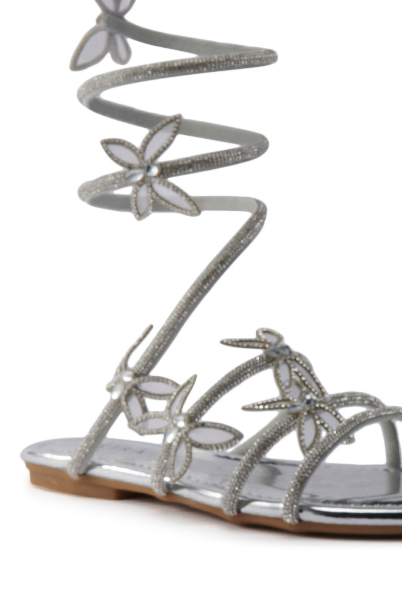 The AKIRA ALBUS SILVER COIL RHINESTONE FLAT SANDAL features metallic faux leather, spiral rhinestone straps with floral accents, wraps elegantly around the ankle, and boasts a flat sole with a shiny silver finish. 6