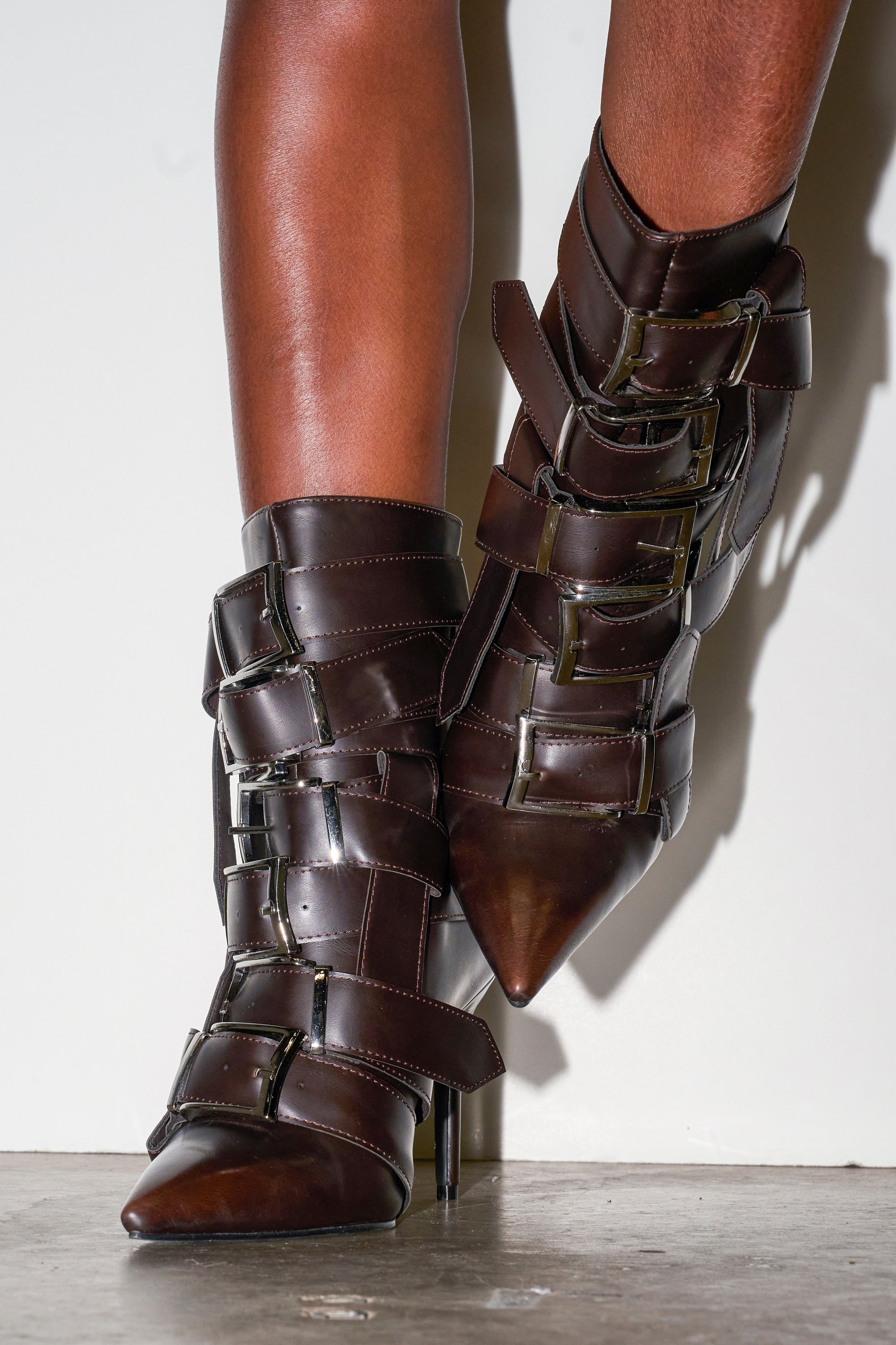 A pair of legs wears the AZALEA WANG ALCETAS CHOCOLATE STILETTO BOOTIE WITH BUCKLES—dark brown, pointed-toe booties with wide buckled straps—standing against a white wall and concrete floor.