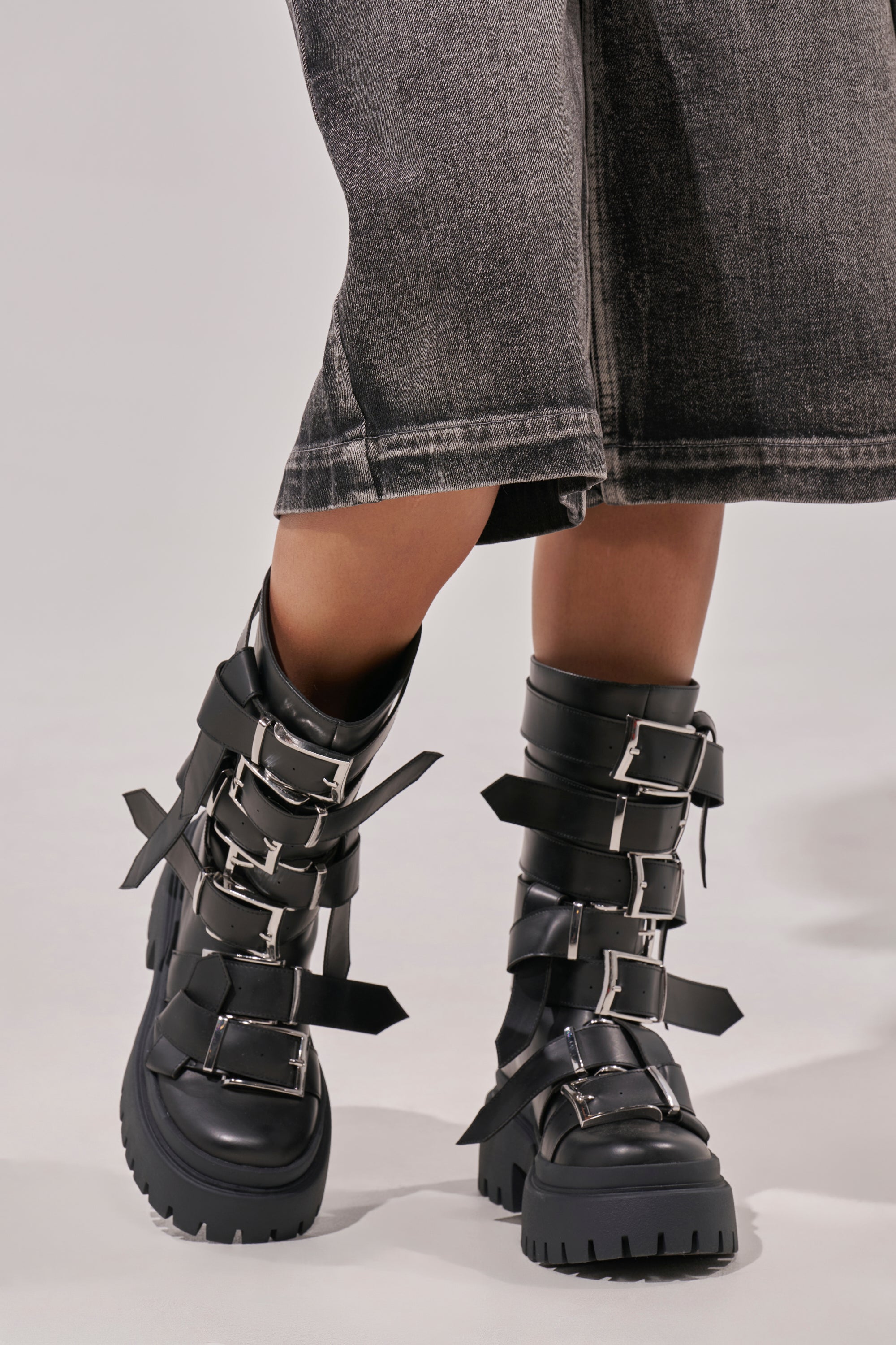 Someone wearing the AZALEA WANG ALCETASA Black Combat Bootie with buckle detail, featuring chunky heels and multiple silver straps, paired with a faded black denim skirt falling below the knees. 1