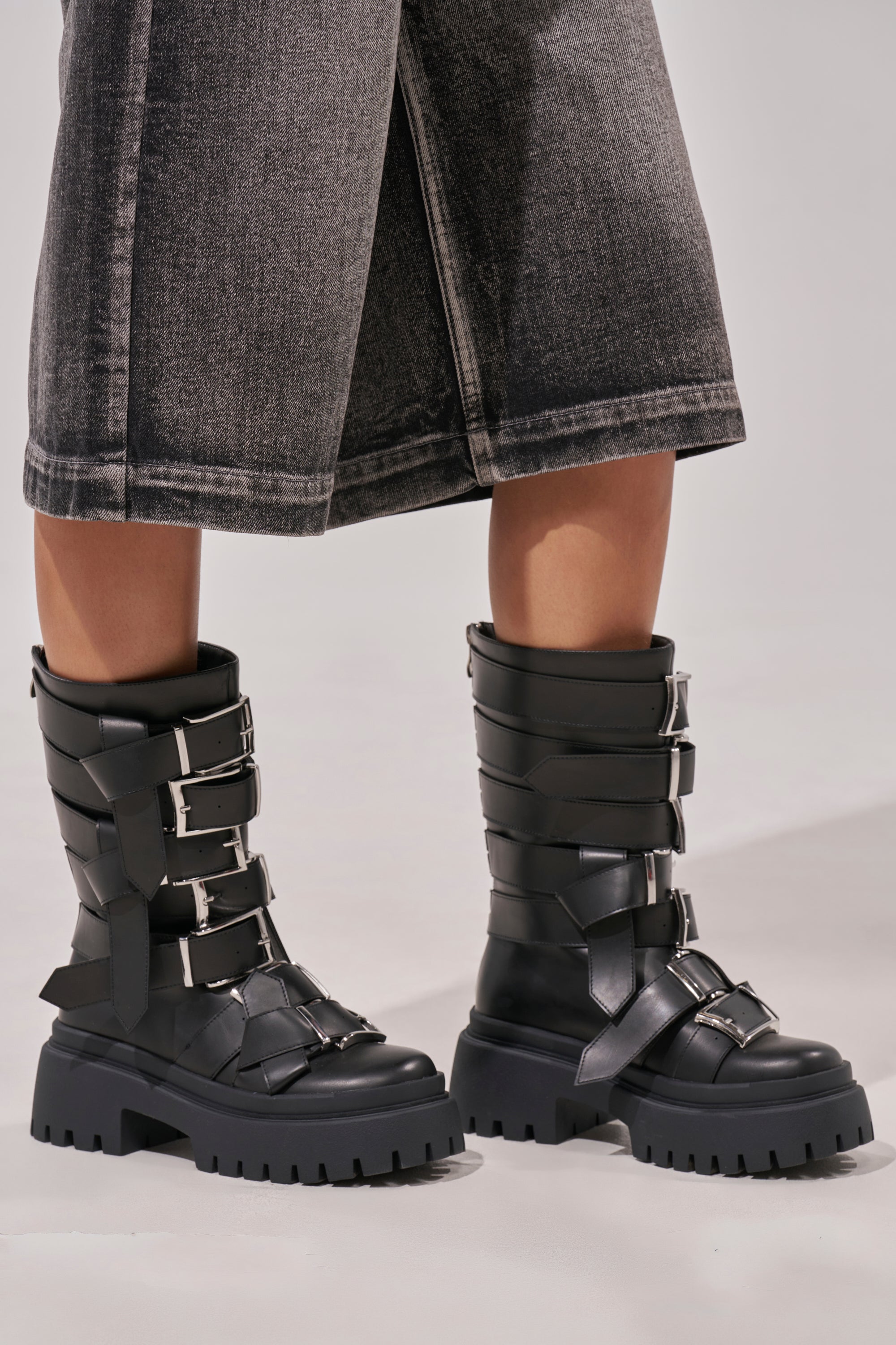 Wearing AZALEA WANG ALCETASA BLACK COMBAT BOOTIE WITH BUCKLE DETAIL and wide-leg, faded black denim culottes, a person stands on a neutral background. 2