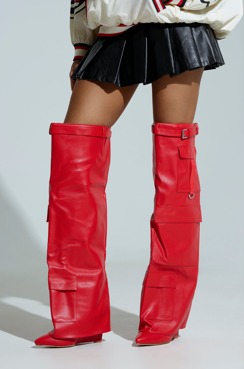 A person is flaunting the AZLAEA WANG ALEXIA RED WEDGE BOOTS, which are sleek, tall faux leather with buckles and large pockets, paired with a black pleated skirt. The shiny red thigh-highs stand out against a plain, light backdrop.