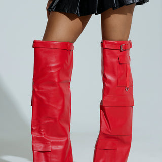 A person is flaunting the AZLAEA WANG ALEXIA RED WEDGE BOOTS, which are sleek, tall faux leather with buckles and large pockets, paired with a black pleated skirt. The shiny red thigh-highs stand out against a plain, light backdrop.