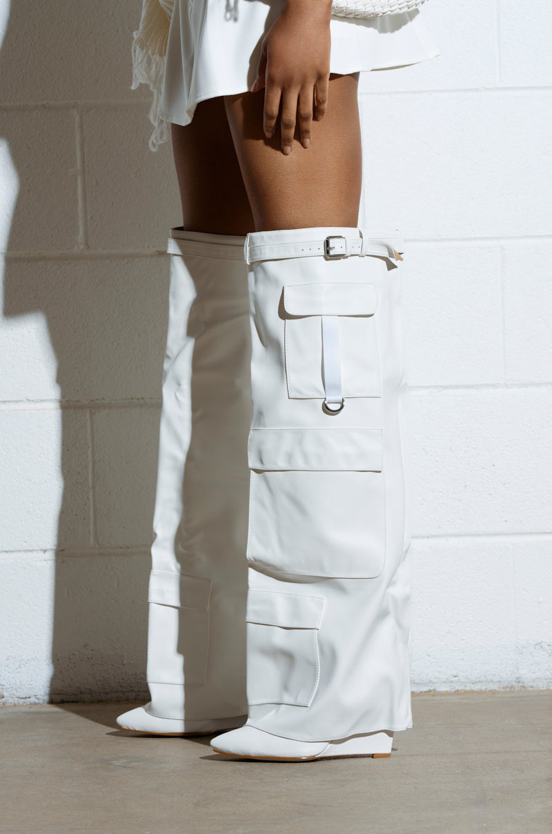 Against a white brick wall, a person in oversized white pants with large pockets and buckles showcases chic faux leather boots. The AZALEA WANG ALEXIA WHITE WEDGE BOOT peeks from beneath the pants, revealing its stylish design as a ruffled white skirt adds texture to the outfit. 0