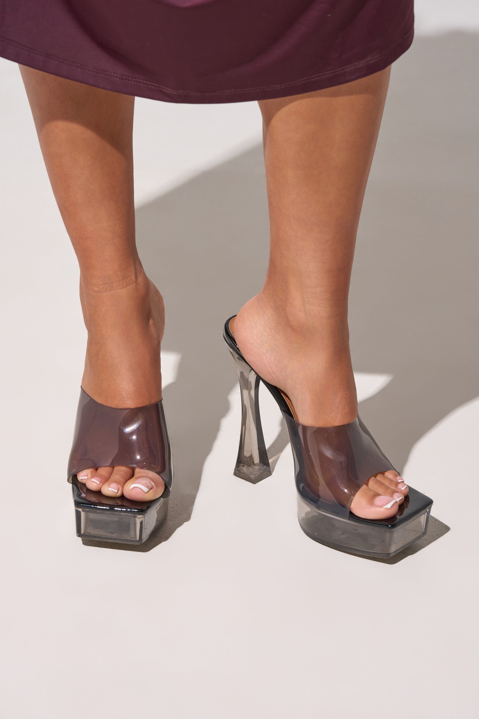 Someone wearing the AKIRA ALEXUS SMOKE PVC CHUNKY SLIP ON SANDAL, which features clear straps, a thick angled chunky heel, and a square platform, stands on a light surface with pale pink painted toenails. 0