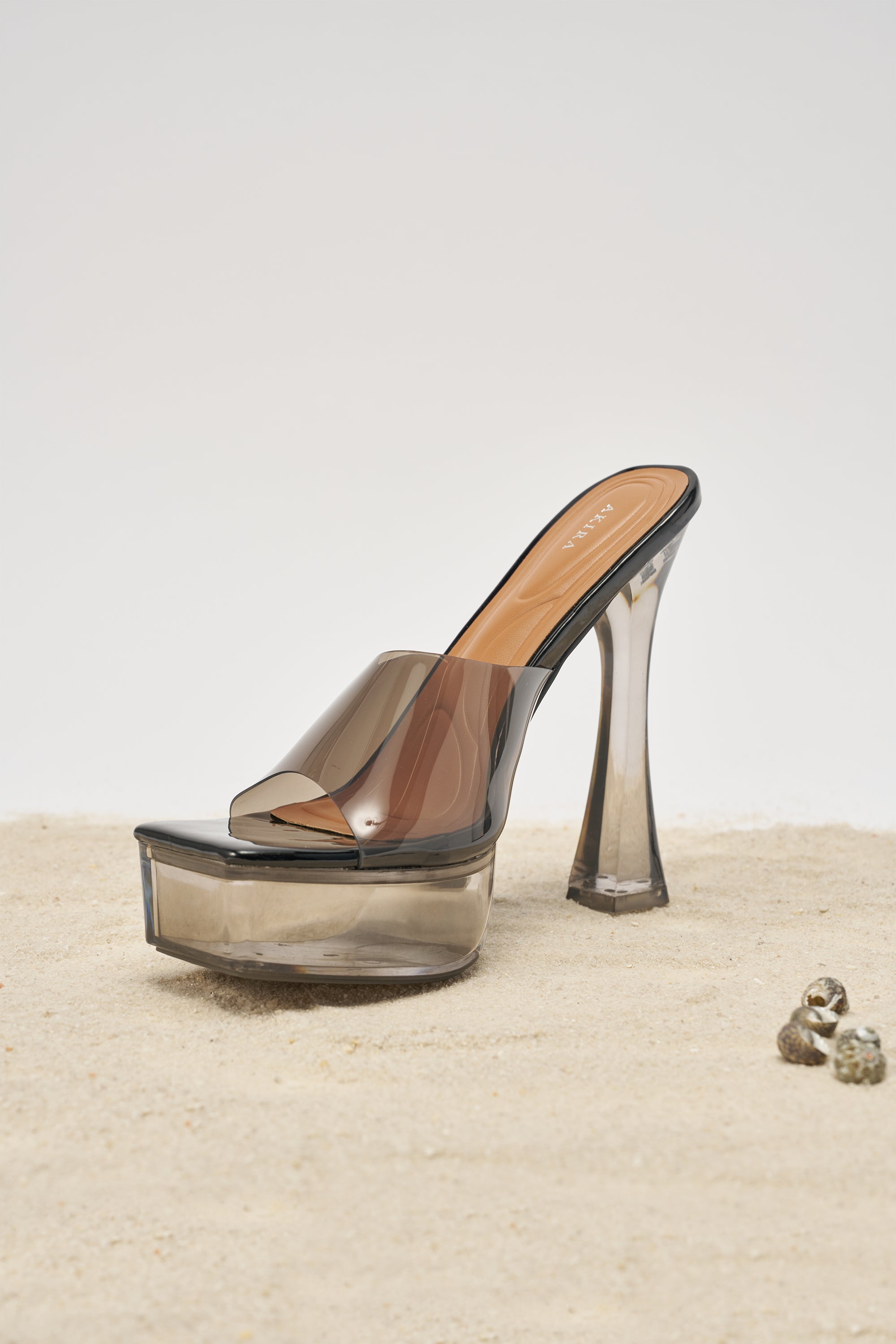 The AKIRA ALEXUS SMOKE PVC CHUNKY SLIP ON SANDAL, featuring a brown-toned upper and clear platform heels, is displayed on sandy ground with small seashells nearby. 3