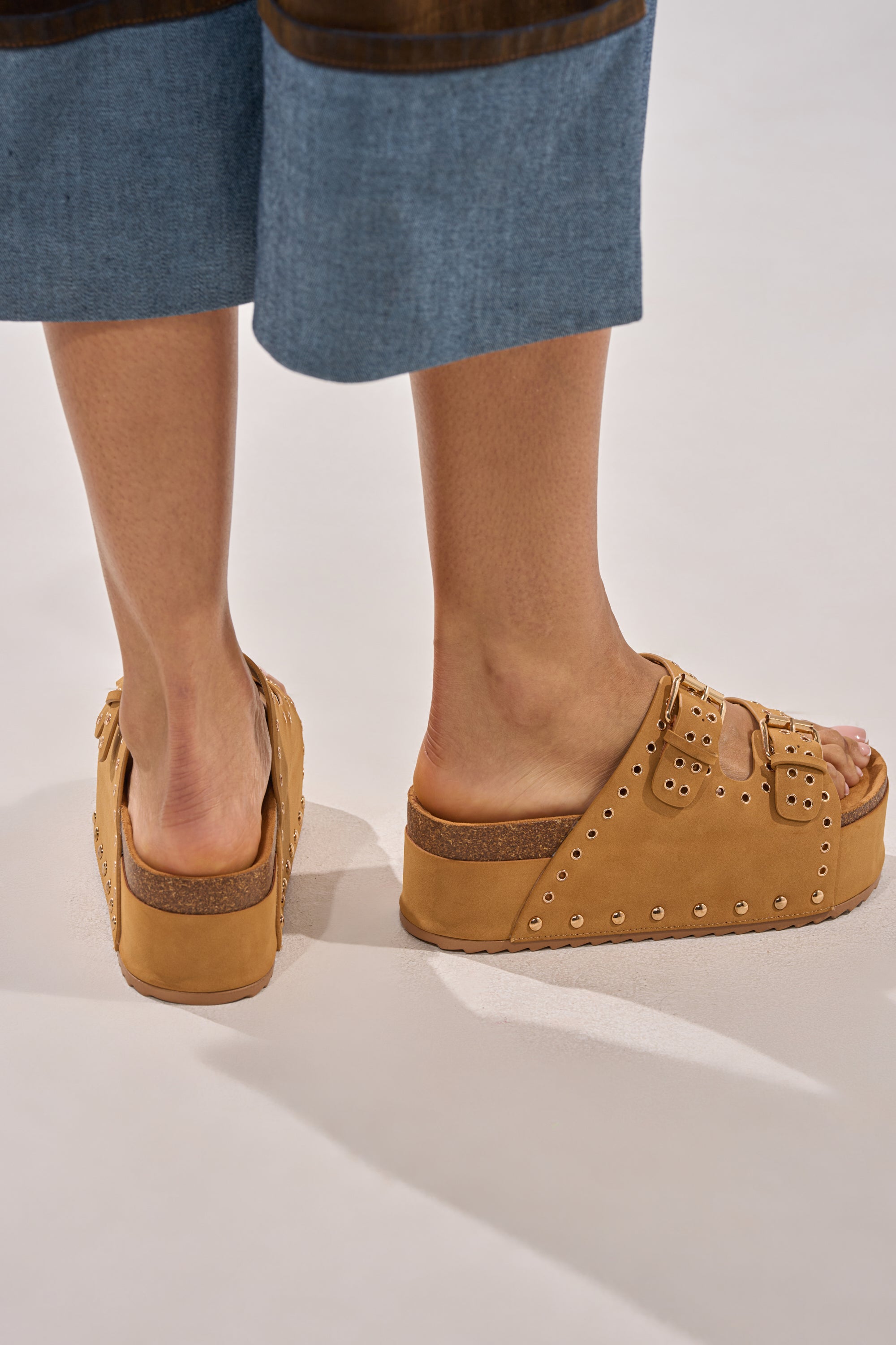 A person in knee-length blue denim shorts wears the AZALEA WANG ALGEDI TAN PLATFORM SANDAL, featuring chunky platforms with metal studs and buckles, while standing on a light-colored surface. 2