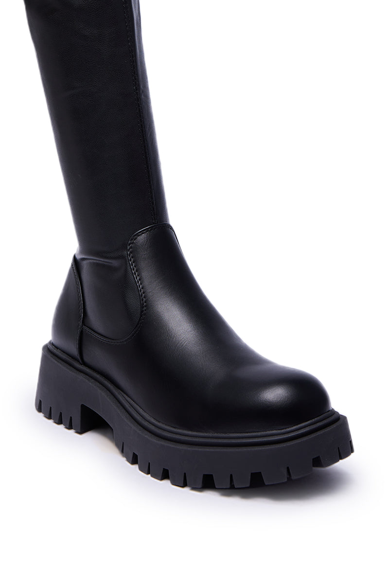 A close-up of the AZALEA WANG ALINAFE BLACK OVER THE KNEE STRETCH PU BOOT shows its black faux leather, chunky treaded sole, and over-the-knee design against a white background.