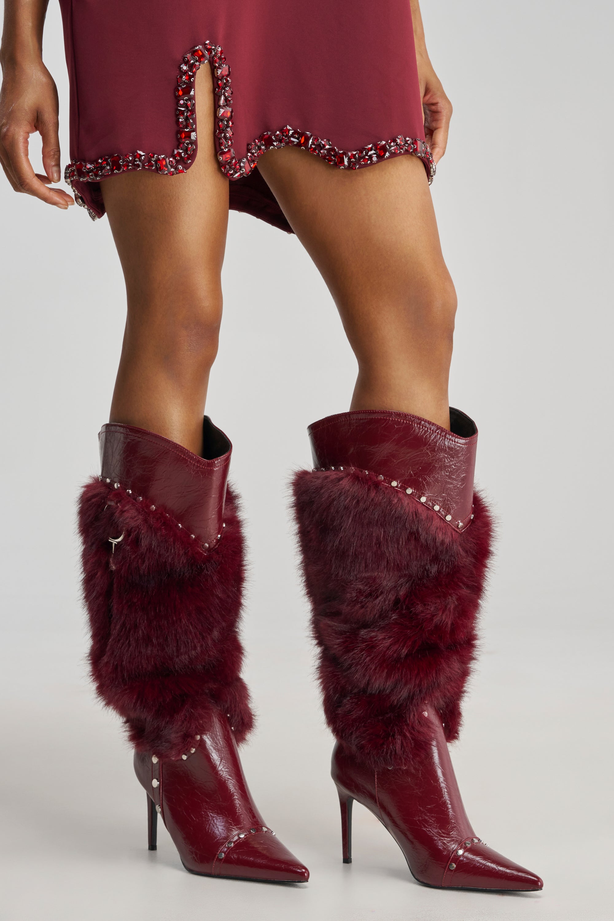 A person is shown, cropped above the knees, wearing tall pointed knee-high AZALEA WANG ALISETTE Burgundy Mixed Material Fur and Faux Leather Stiletto Boots featuring studded details and faux fur accents, along with a burgundy dress with a front slit and decorative trim.