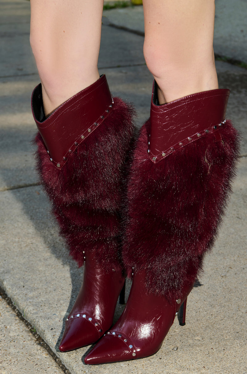 A close-up of a person wearing AZALEA WANG ALISETTE Burgundy Mixed Material Fur and Snake Stiletto Boots with pointed toes, fur and snake-textured accents, standing on a concrete sidewalk.