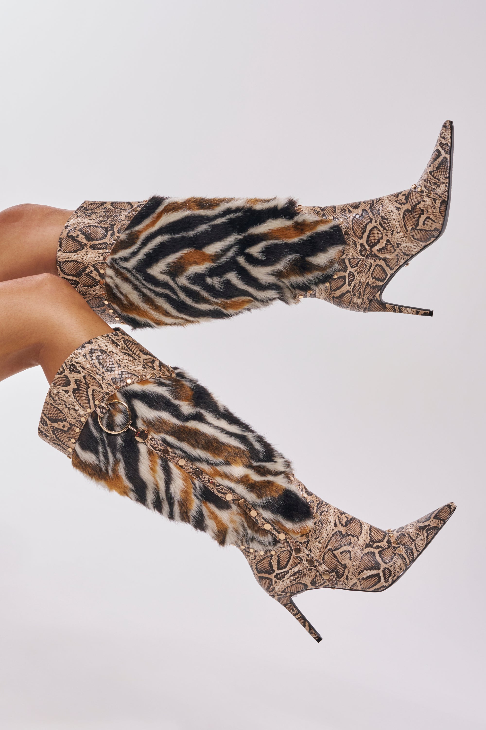 The AZALEA WANG ALISETTE SNAKE MULTI FUR STILETTO BOOT features pointed toes with faux snakeskin and animal fur patterns in brown, black, beige, and white, shown on crossed legs against a plain background.