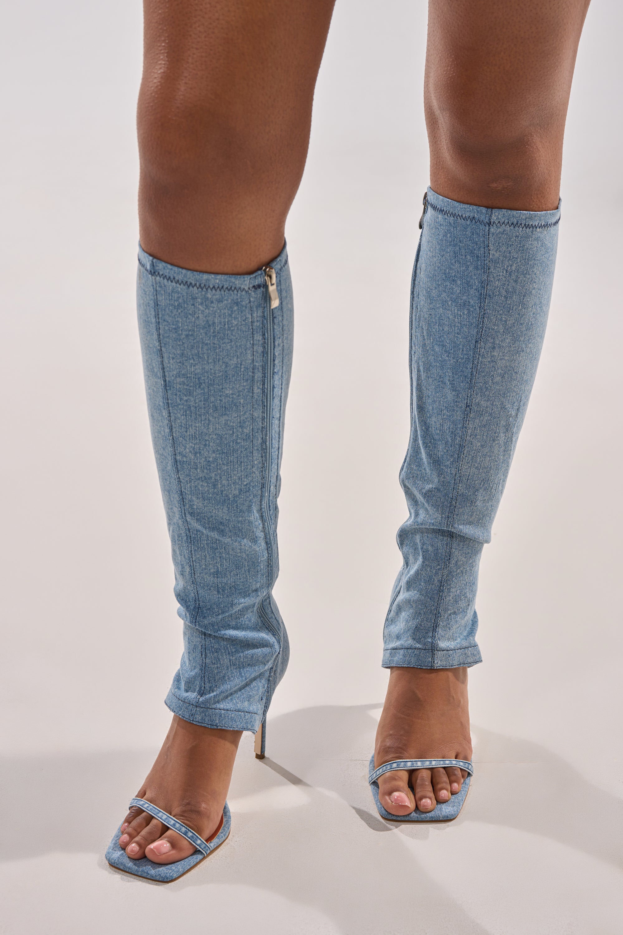 A person stands confidently on a light surface wearing the AZALEA WANG ALISHAS DENIM GLADIATOR STILETTO SANDAL—stylish open-toe denim heels with zippers and stiletto heels, paired with matching high heel sandals.
