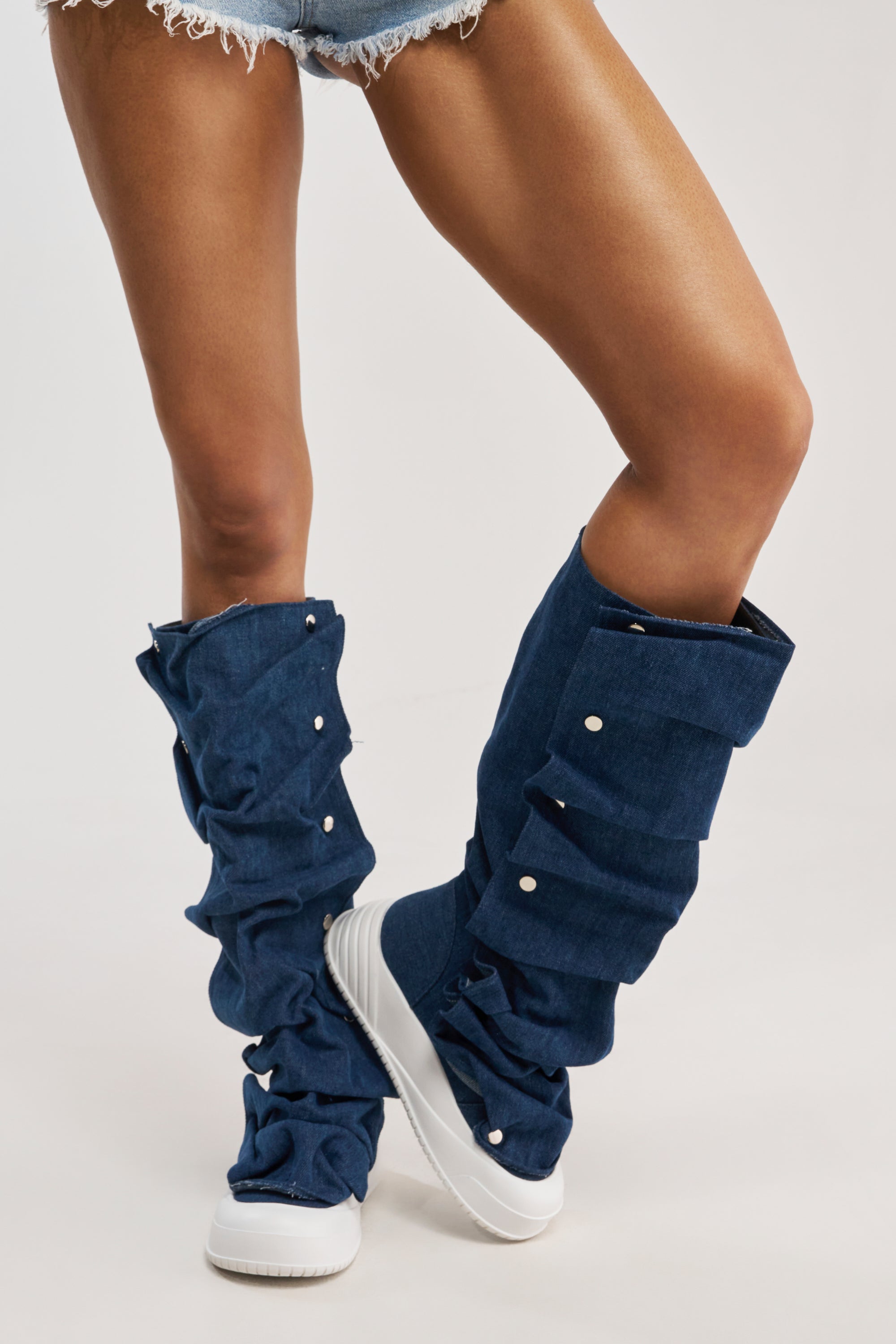 A person wears AKIRA GOLD ALIXY DENIM FLAT RUFFLE BOOTS with frayed denim shorts and white sneakers, creating a casual streetwear vibe with the unique slouchy leg covers and white buttons against a plain background.