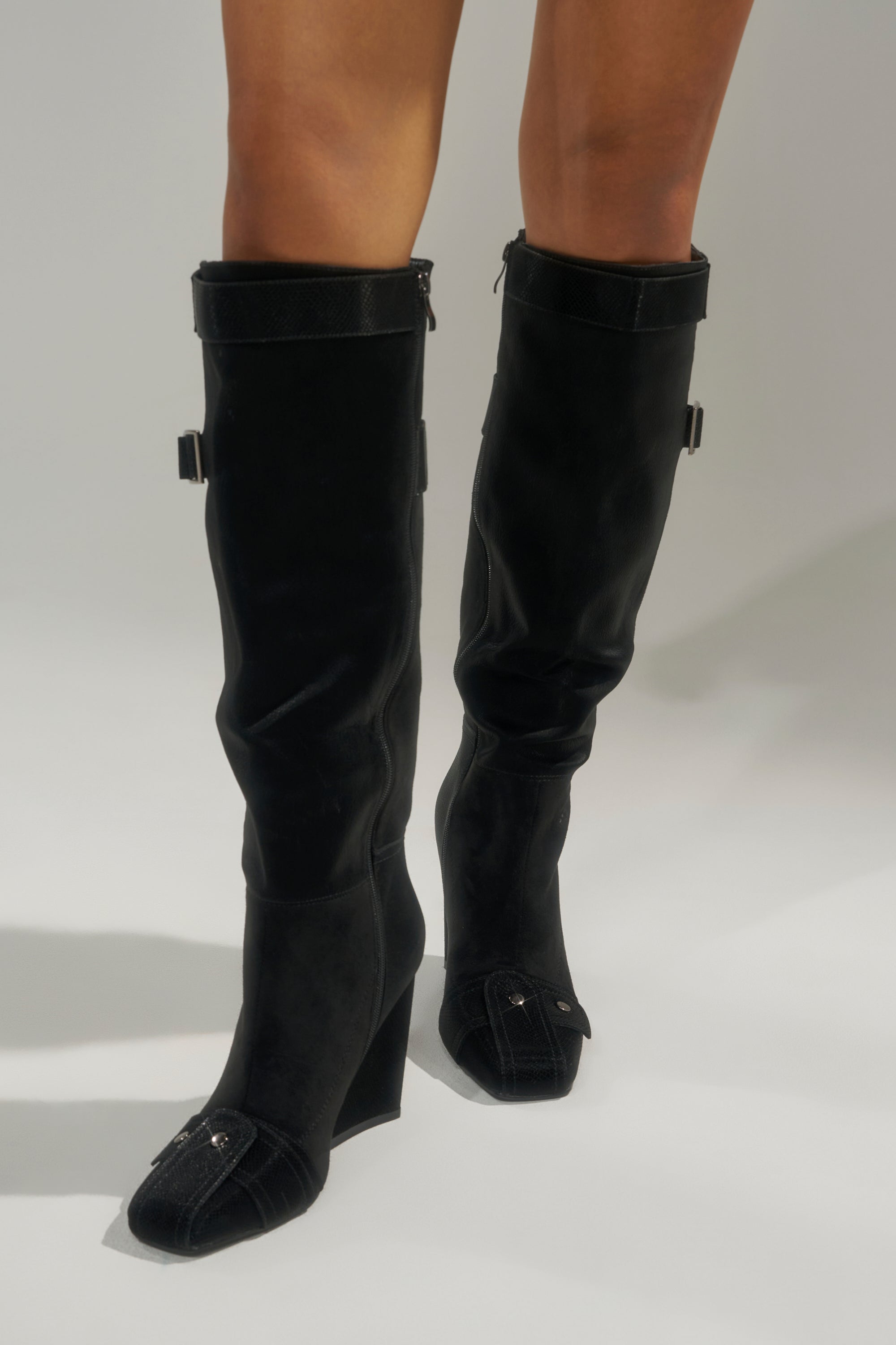 Someone wearing the AZALEA WANG ALLONAS BLACK KNEE HIGH CHUNKY BOOT, featuring side buckles and decorative flaps with button details at the square toe, stands on a light-colored surface.