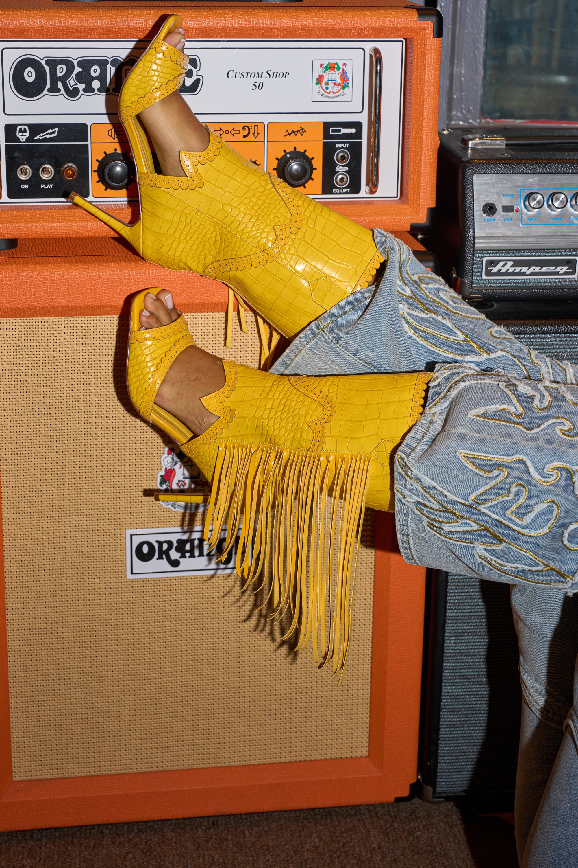 A person wearing the AZALEA WANG ALPHONSE YELLOW OPEN TOE SANDAL and embroidered jeans rests their feet on an orange guitar amplifier, with another amp seen in the background. 0