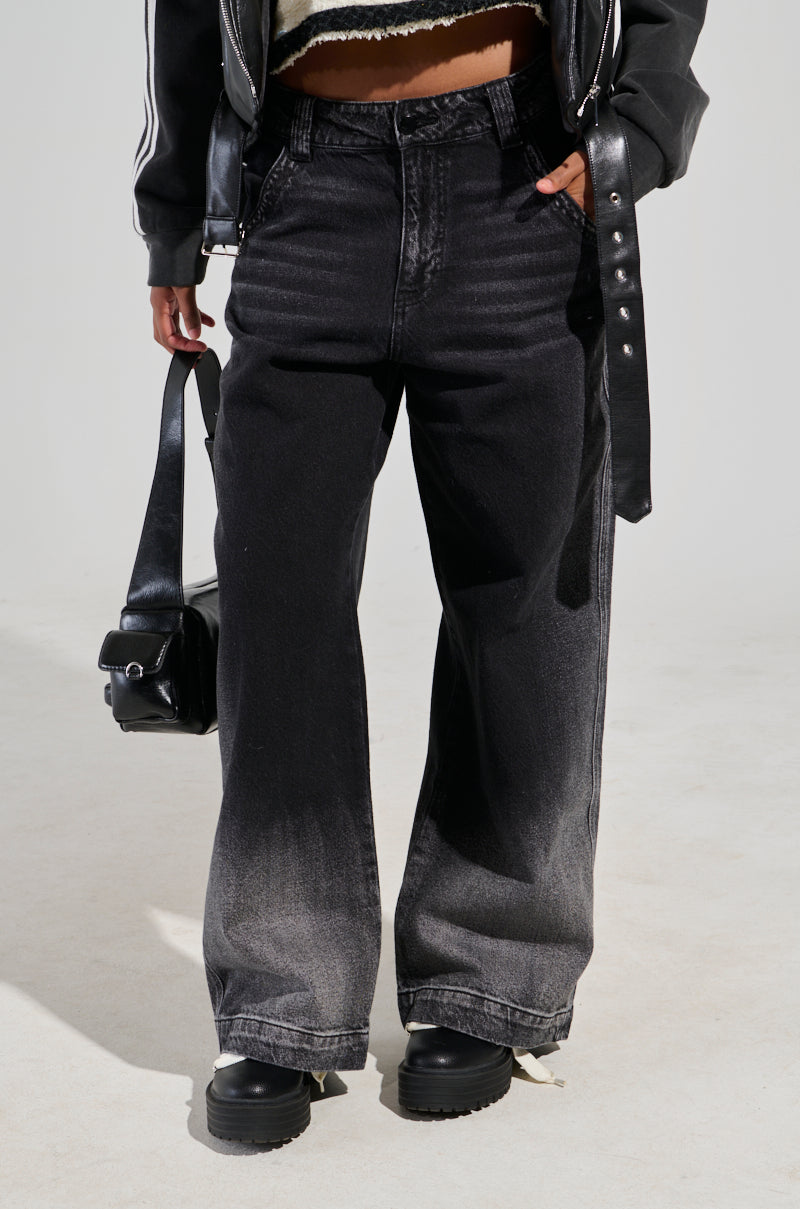 A person wearing the "THERES NO OTHER BLACK DENIM PANT" featuring a high-rise, wide-leg silhouette and a black leather jacket holds a sleek black bag. The ensemble is completed with stylish black platform shoes as the person stands against a light gray background.