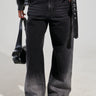 A person wearing the "THERES NO OTHER BLACK DENIM PANT" featuring a high-rise, wide-leg silhouette and a black leather jacket holds a sleek black bag. The ensemble is completed with stylish black platform shoes as the person stands against a light gray background.