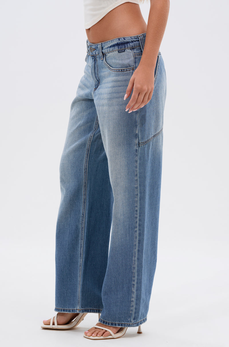 A person stands against a plain white background wearing RELAX WITH ME WIDE LEG DENIM in light blue vintage wash, paired with a white crop top and white heeled sandals.