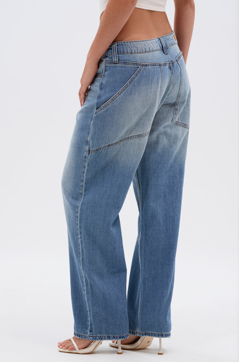 A person wearing RELAX WITH ME WIDE LEG DENIM, featuring a vintage wash and diagonal seam details, styled with a white cropped top and heeled sandals, stands against a plain white background.