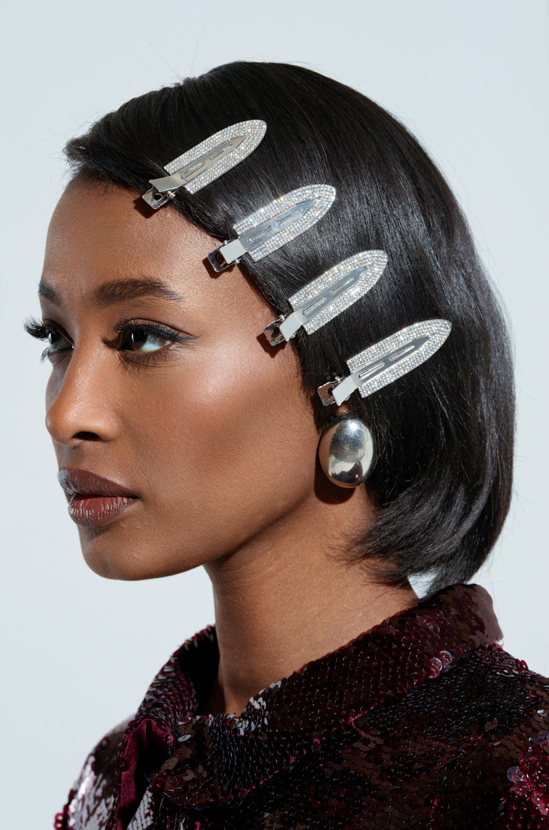 A person with sleek, short hair adorned with the "TOO PRETTY FOR THIS BLING HAIR CLIPS," wearing bold metallic earrings and a textured, dark-colored top. They have elegant makeup, including winged eyeliner and glossy lipstick. 1