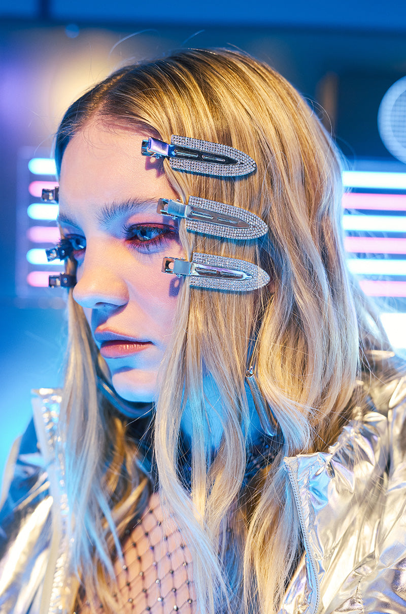 Someone with long blonde hair showcases the "TOO PRETTY FOR THIS BLING HAIR CLIPS" while wearing a silver metallic jacket. Rhinestone embellishments amplify the sparkle, set against a backdrop of blurred neon lights, creating a futuristic and colorful ambiance. 0