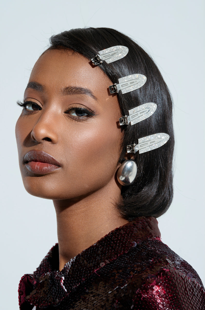 A woman with sleek hair wears large TOO PRETTY FOR THIS BLING hair clips and a silver earring, paired with a shiny, textured top. Her expression is calm, with neutral makeup accentuating her features against a light background. 2