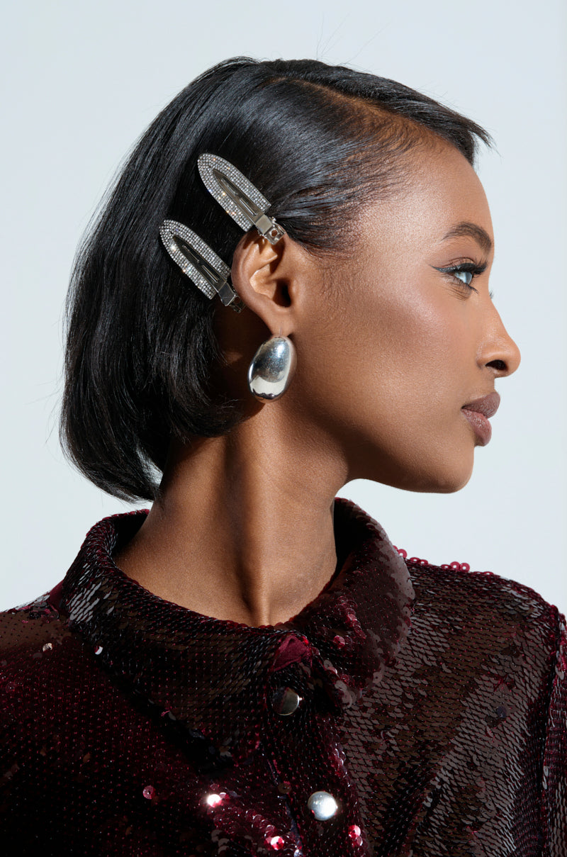 A person with short black hair is shown in profile, adorned with TOO PRETTY FOR THIS BLING HAIR CLIPS and large, oval earrings. They are dressed in a burgundy sequined top against a plain background. 3