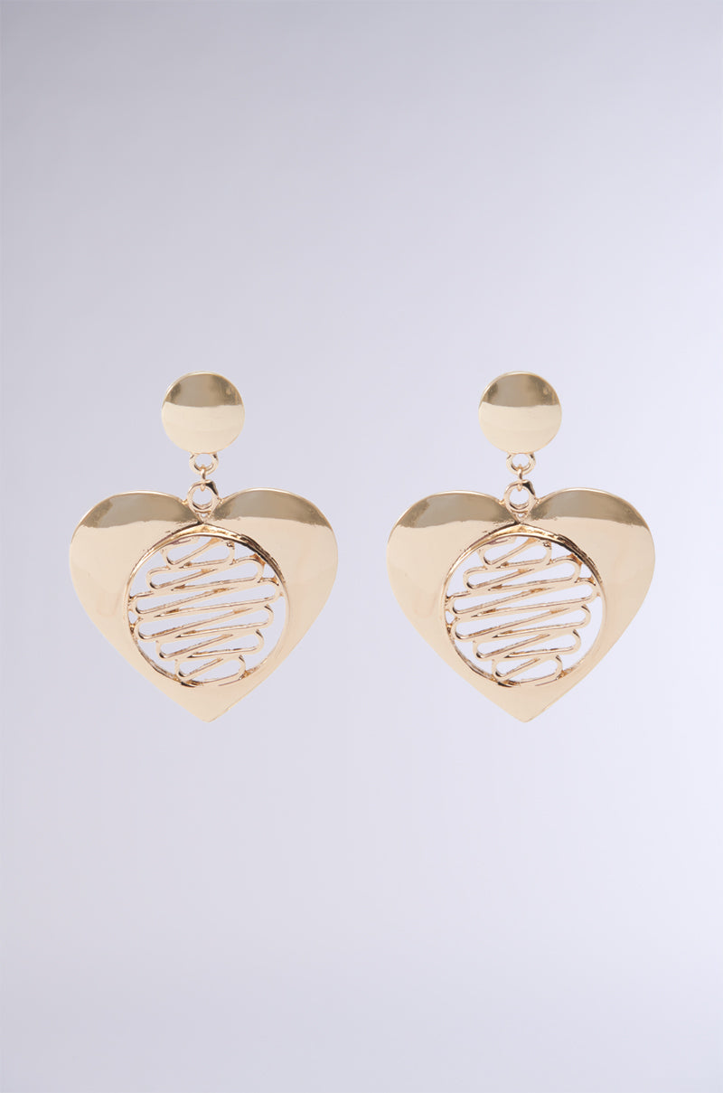 The TIE MY HEART EARRING features a pair of heart-shaped faux gold earrings with a circular, intricate wire pattern in the center, attached to a small gold circular stud. The metallic detailing stands out beautifully against a plain, light gradient background from white to light gray.