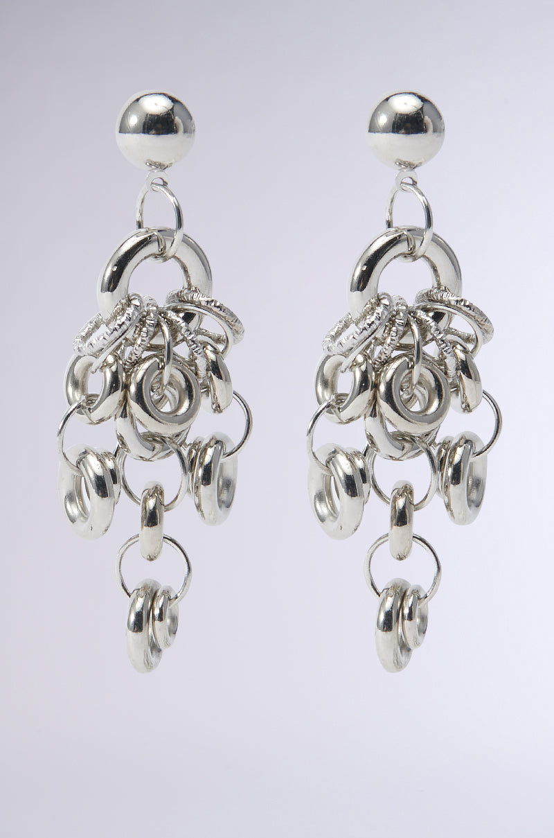Introducing the MODERN MUSE EARRING: a pair of faux silver dangling earrings featuring an intricate arrangement of interlocking circular loops and O-ring attachments, all suspended from a round stud. The varying sizes of the loops create a cascading effect with their shiny, metallic finish against a light background.