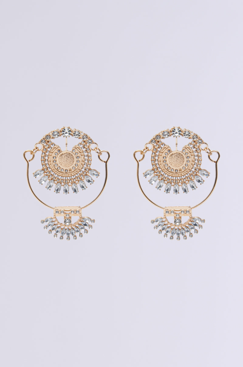 The ART DECO DREAMING EARRING features elegant gold circles, reminiscent of vintage luxury, set with sparkling clear gemstones and dangling rhinestones. The intricate design creates a sophisticated look against a soft gray background.
