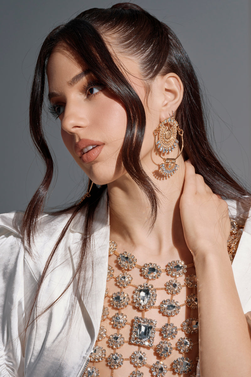 A woman with long brown hair styled in loose waves showcases the ART DECO DREAMING EARRING, pairing it with a jeweled top and white satin garment for a vintage luxury look against a plain background.