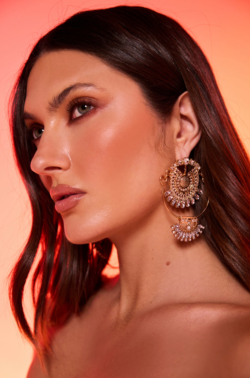 A woman with long, wavy brown hair and glowing skin poses against a warm orange gradient, showcasing the ART DECO DREAMING EARRING—vintage luxury earrings adorned with crystals—while wearing natural makeup.