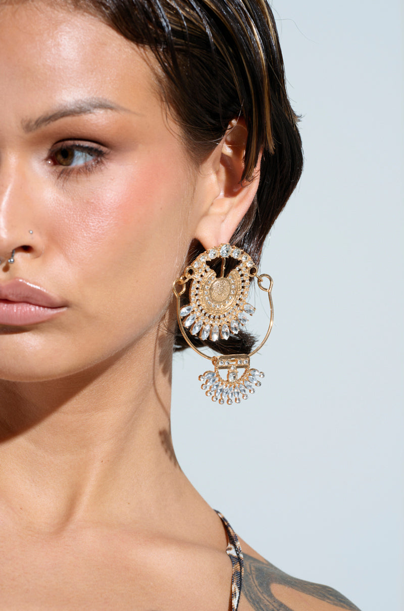 A person with wet, short hair wears ART DECO DREAMING EARRINGS adorned with crystals. They also have a facial piercing and a subtle tattoo on their shoulder. The light, solid color background enhances the vintage luxury vibe.