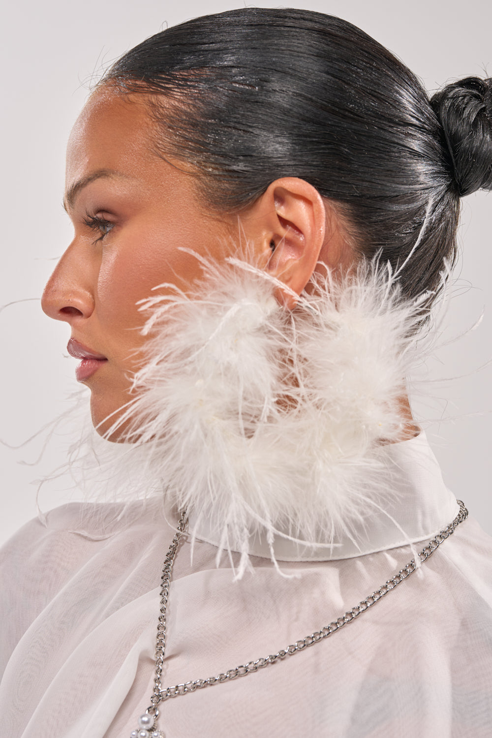 A woman with slicked-back dark hair wears LET ME FLY HOOP EARRING—large white feather-lined hoops—with a sheer white top and silver chain necklace, shown in profile against a plain background. 0