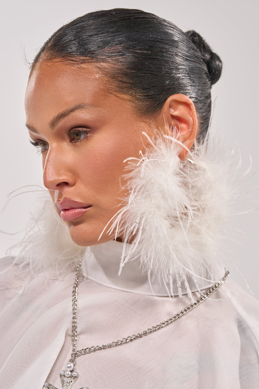 A woman with sleek, dark hair in a bun wears the LET ME FLY HOOP EARRING, a sheer white top, and a silver chain necklace. She gazes calmly to the side against a plain background. 1