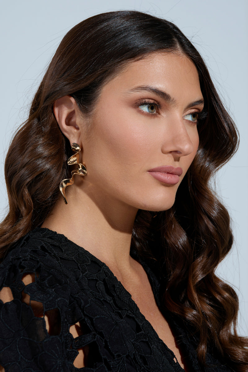 A woman with long, wavy brown hair wears a black lace top and large Gold Squiggles Dangle Earrings. Her neutral expression complements her abstract silhouette as she gazes to the side against a light background, embodying a subtle ribbon-like design in her posture. 1