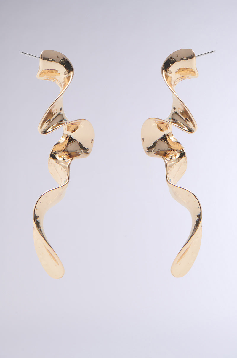 The GOLD SQUIGGLES DANGLE EARRINGS feature an abstract, ribbon-like silhouette with wavy, twisted curves for a modern and artistic look, hanging vertically against a plain gray background. 2
