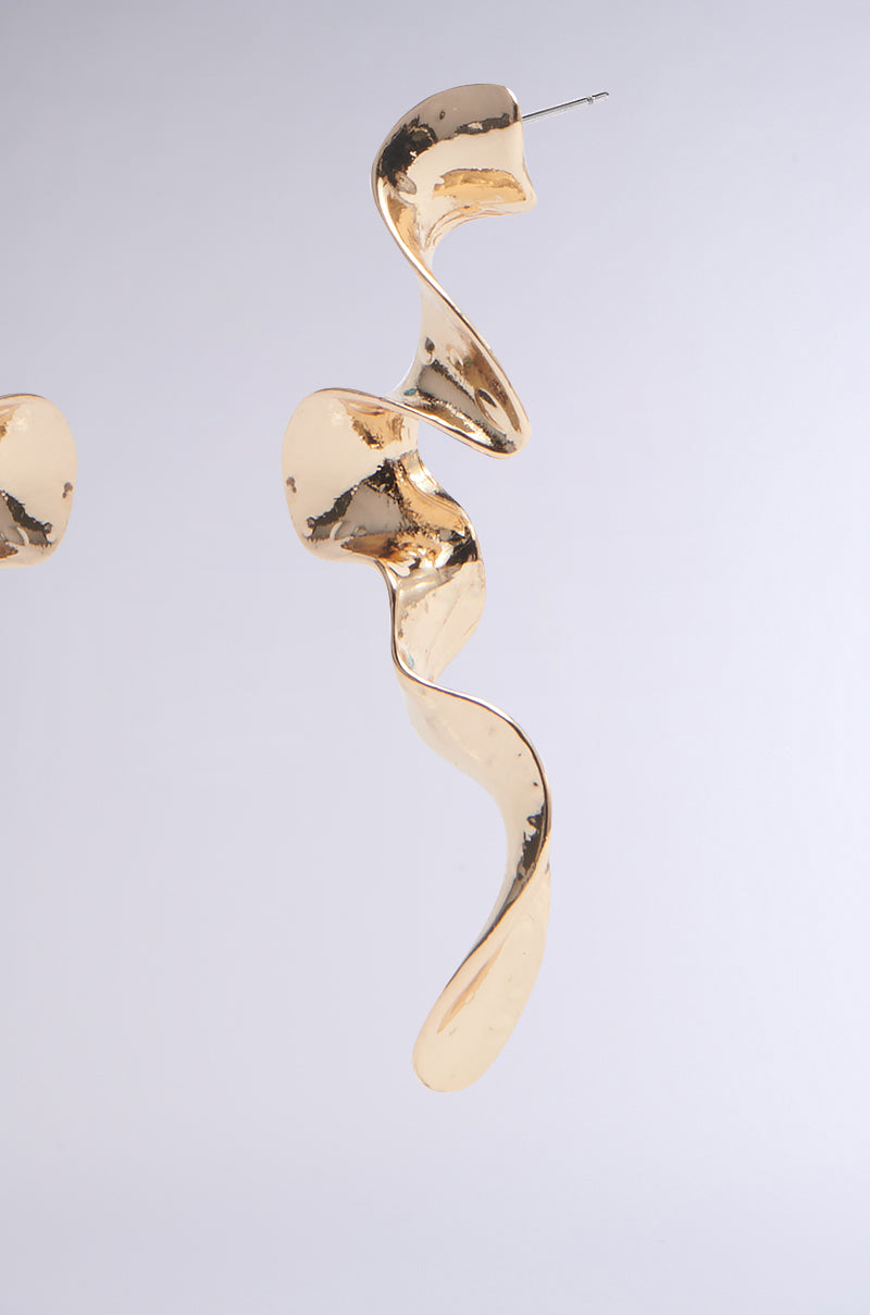 The GOLD SQUIGGLES DANGLE EARRING, with a shiny metallic finish, features an elegant flowing shape and smooth, irregular curves. It is suspended against a light gray background, creating a striking abstract silhouette. 3