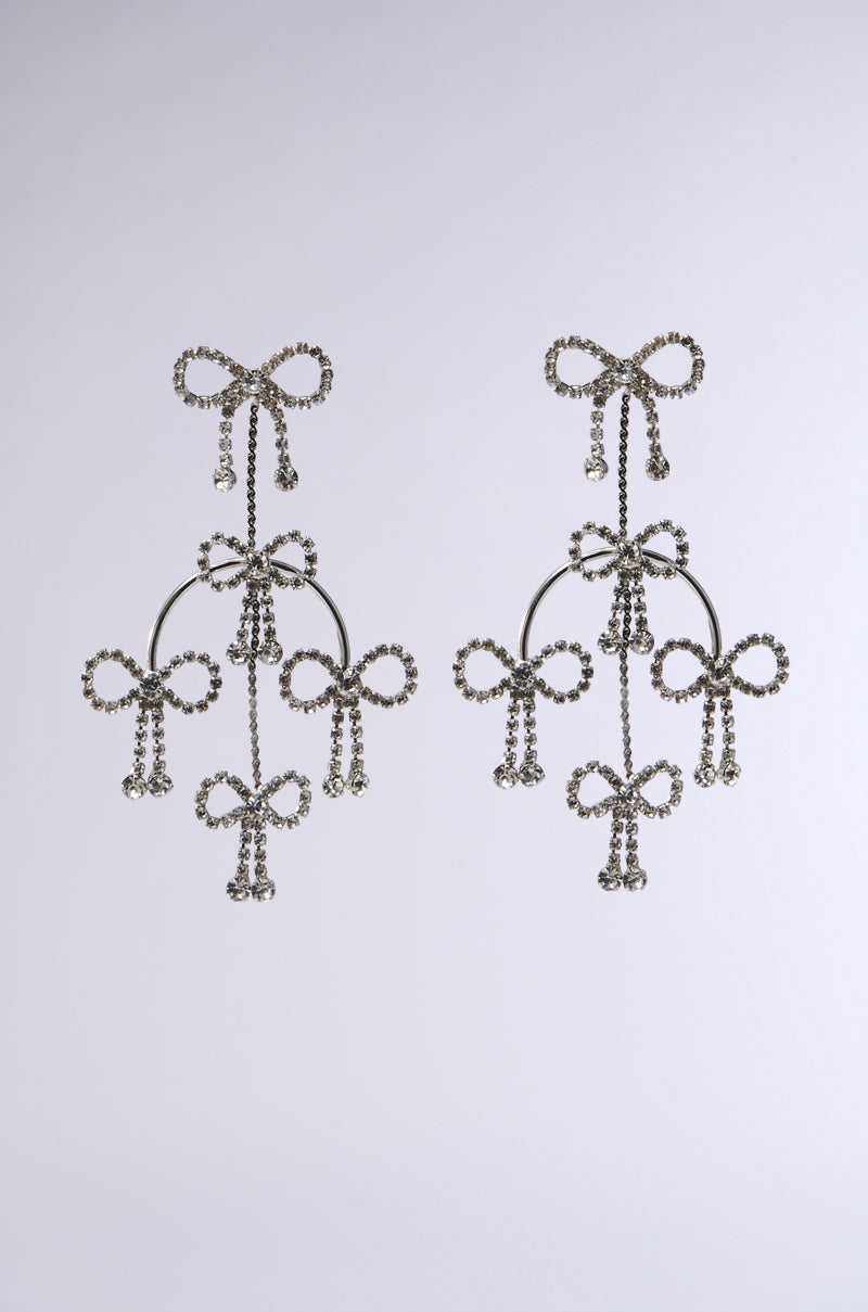The BAD BISHES ONLY EARRING captivates with intricate faux silver metallic bow designs. These chandelier earrings boast rhinestone-adorned bows, elegantly hanging against a plain white background, radiating luxury and charm.
