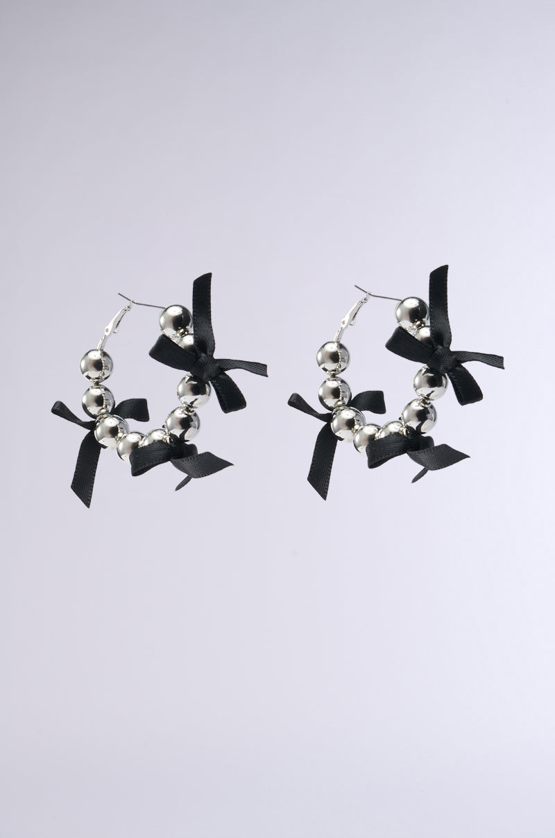 The TIE IT WITH A BOW EARRINGS are silver hoop earrings, beautifully detailed with shiny metallic and silver beads, accented by small satin black bows. Set against a simple grey background, they exude understated sophistication.