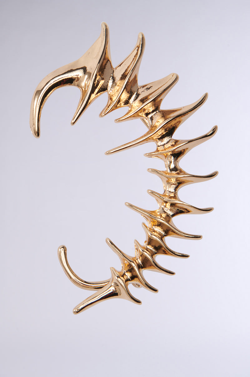 The I BOUNCE BACK EAR CUFF is a large, sculptural piece in faux gold with a curved, spiky spine or claw-like design. Its smooth metallic texture captures the light, showcasing intricate details against a plain gray background. 1