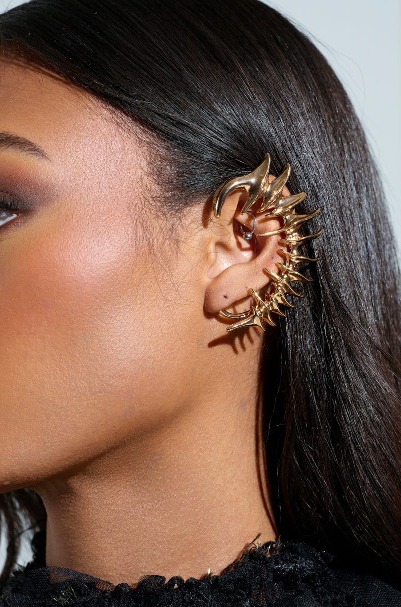 Their right ear features the I BOUNCE BACK EAR CUFF, a striking faux gold accessory with stylized spikes. Straight brown hair complements a black textured garment. 0