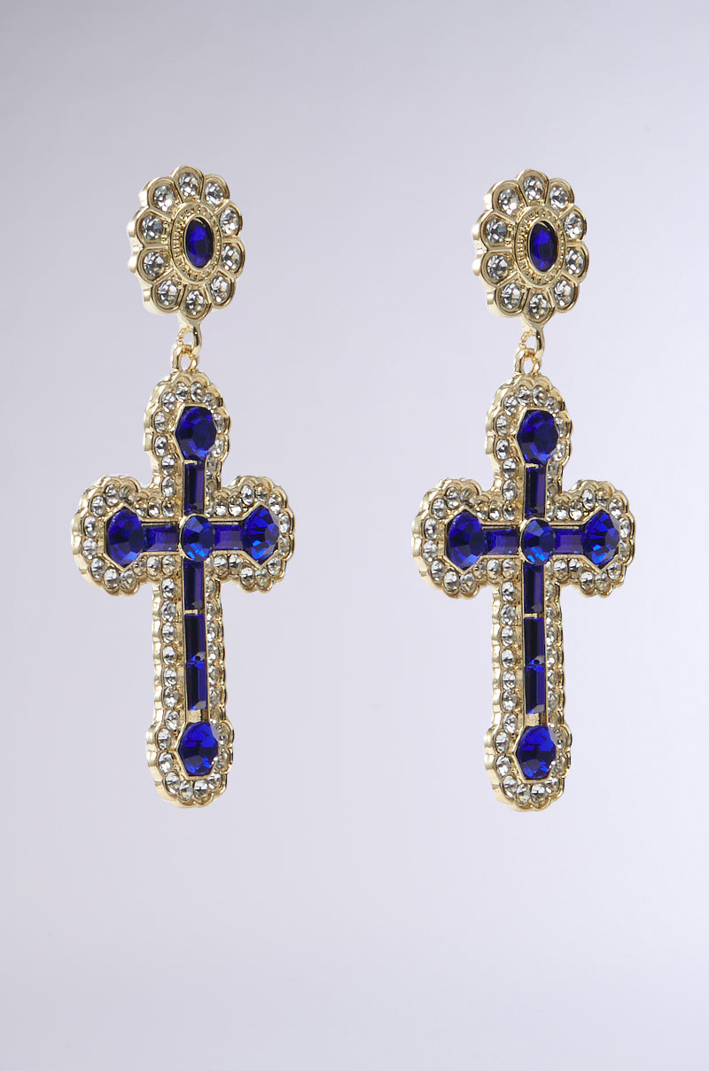 Gold earrings inspired by the QUEEN SHI CROSS EARRING design; they feature blue-jeweled crosses with rhinestone accents and intricate diamond-like detailing, hanging from floral-shaped studs against a plain gray background.