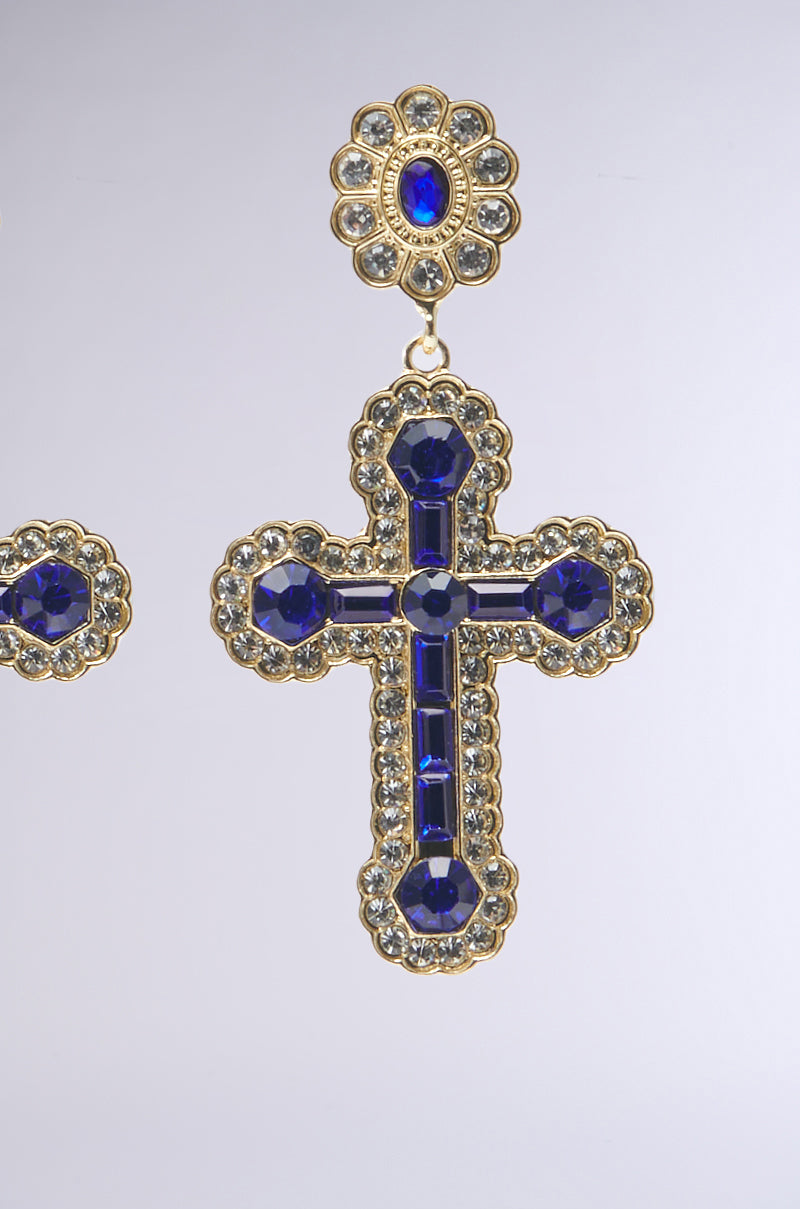 The QUEEN SHI CROSS EARRING is a gold cross design decorated with deep blue faux sapphire jewels and shimmering rhinestones, featuring a floral motif with a central blue gem against an elegant light gray background.
