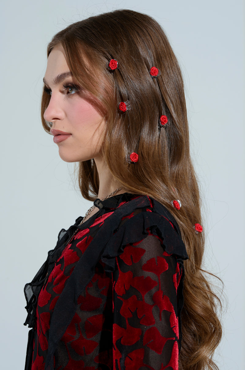 A woman with long brown hair embellished with small red roses and a stylish clip from the ROSES ARE RED HAIR CLIP PACK, wearing a black and red floral dress, faces left. Her makeup features pink blush and subtle eyeshadow against a neutral background.