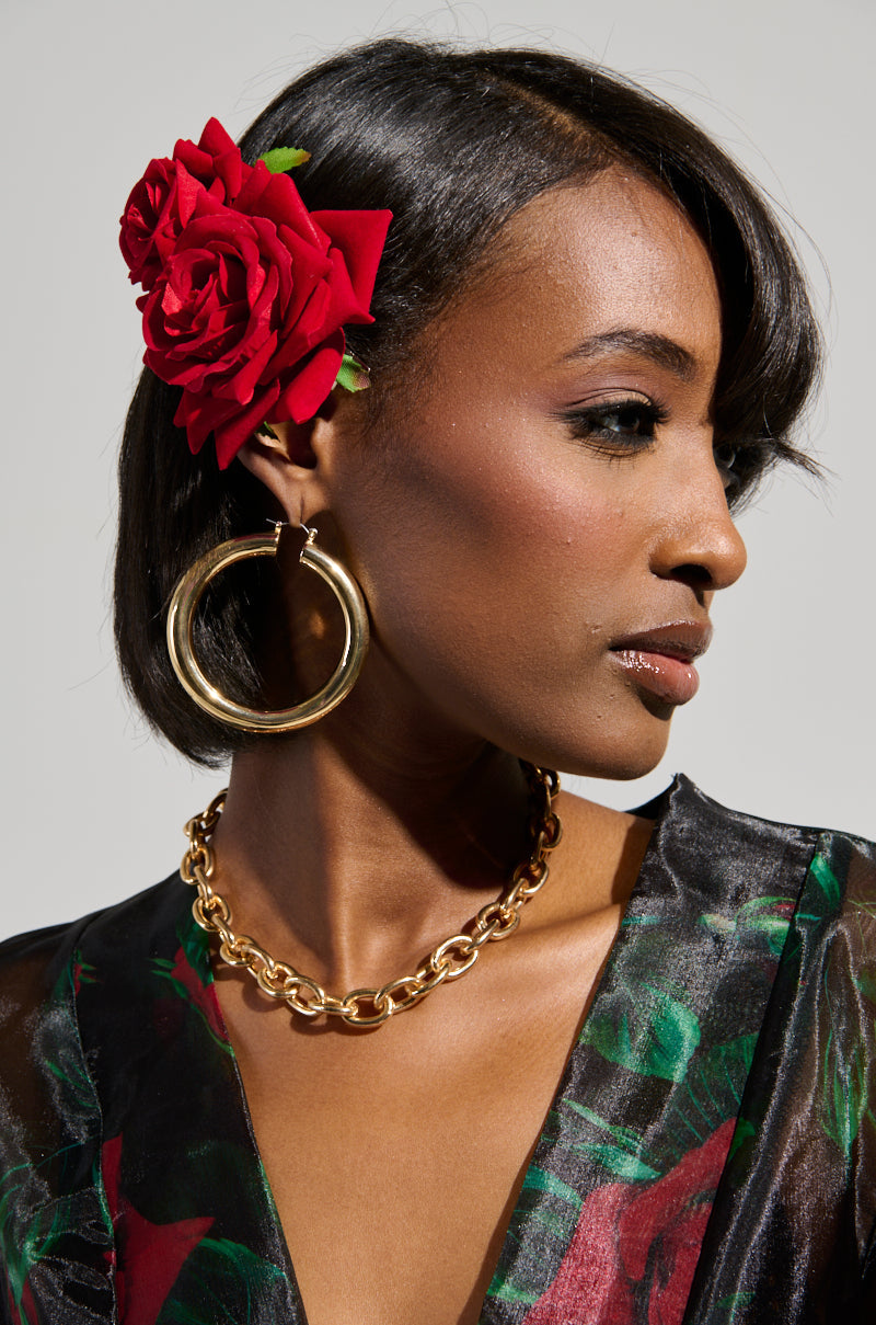 A woman with short black hair accessorized with a DOLCE VITA ROSE HAIR CLIP, complemented by large gold hoop earrings and a gold chain necklace. She is wearing a black and floral ensemble, gazing to the side against a simple backdrop. 0